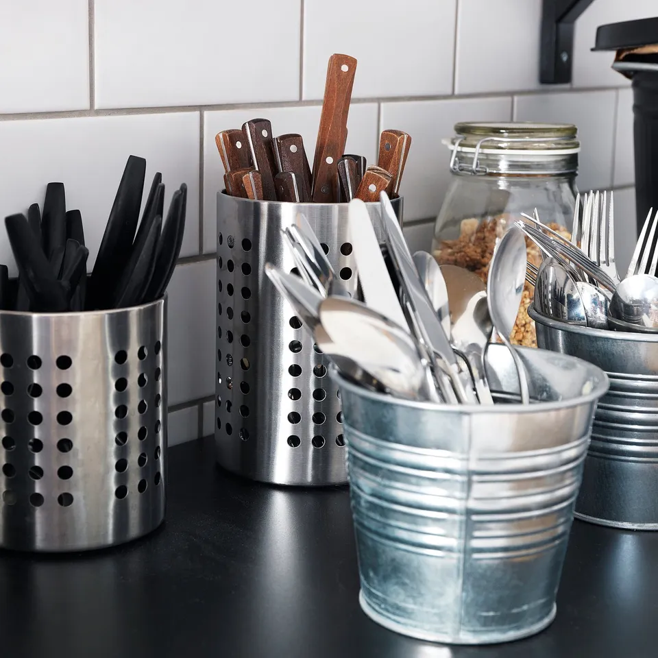 Metal kitchen utensils organised in stainless steel holders on a countertop, with black handles, wooden spoons, and spoons visible.