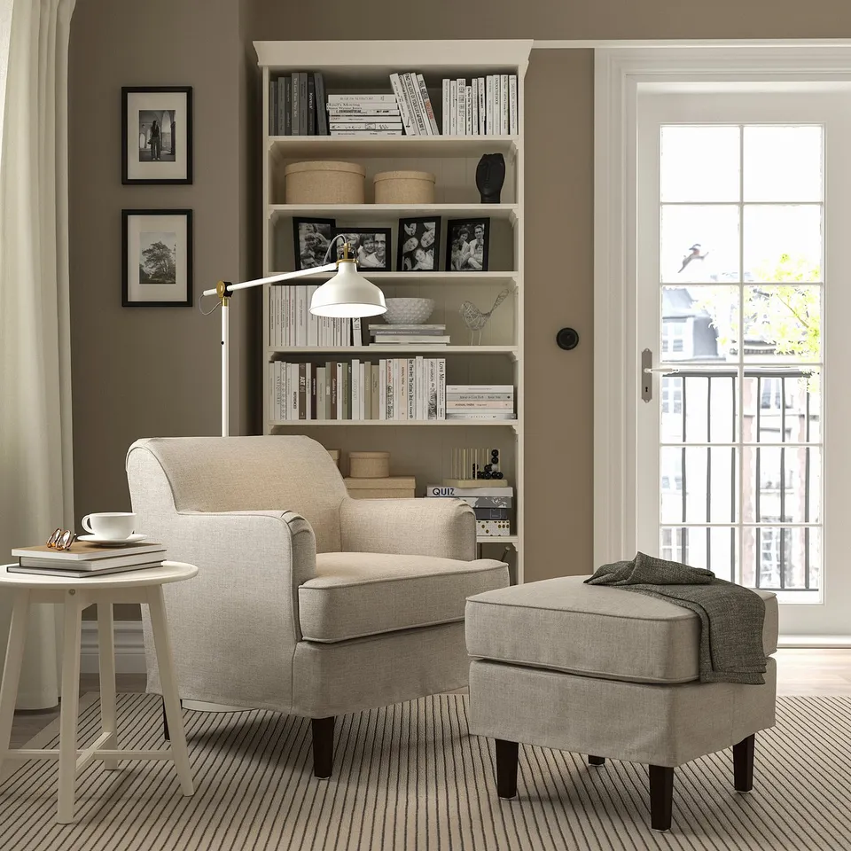 Modern living room: beige rooksjön armchair & ottoman, patterned carpet, bookcase with lamp & decor, large window.