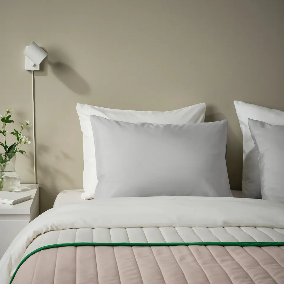 Small, square, white pillow on large, quilted bed, light grey pillowcase, green edge, bedside table with flower vase.