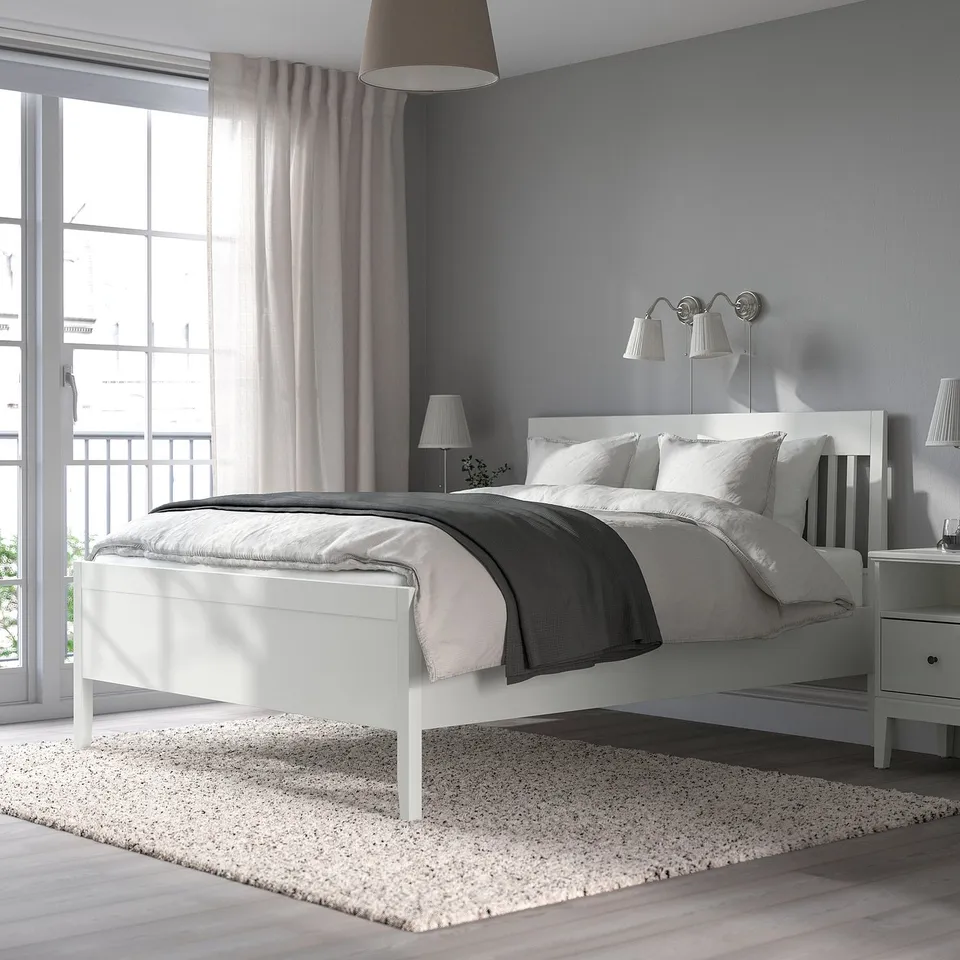 White IDANÄS bed with tall headboard in minimalist room with large window, rug, and pendant lamp.