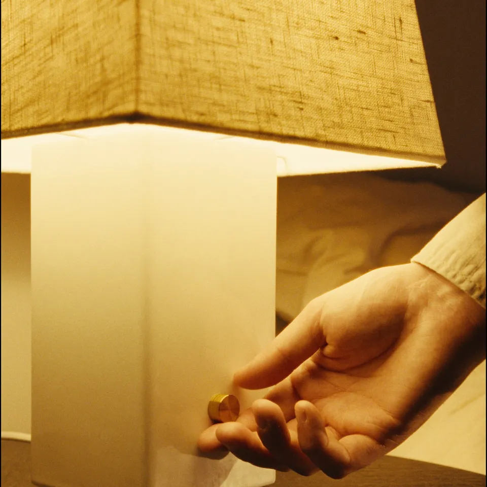 A hand holding a small gold-colored object near a modern lamp with a linen shade and ceramic base.