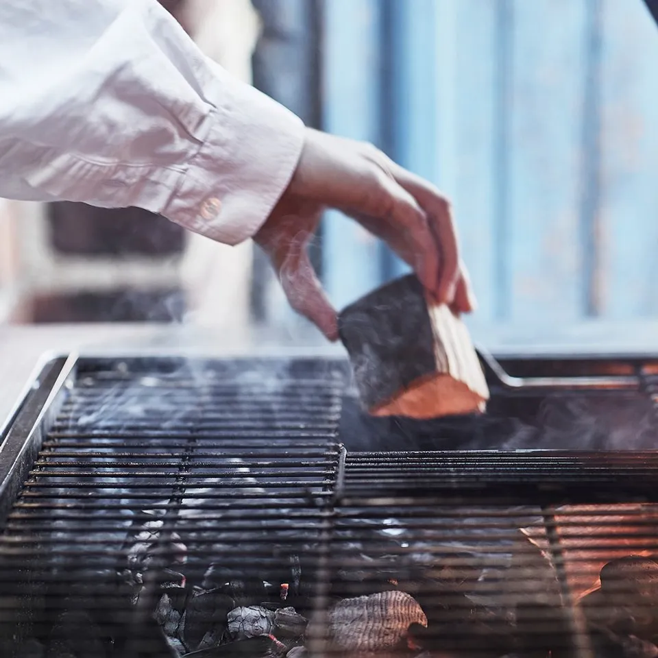 Person grilling on a stainless steel GRILLSKÄR barbecue, using tongs, with flames visible.