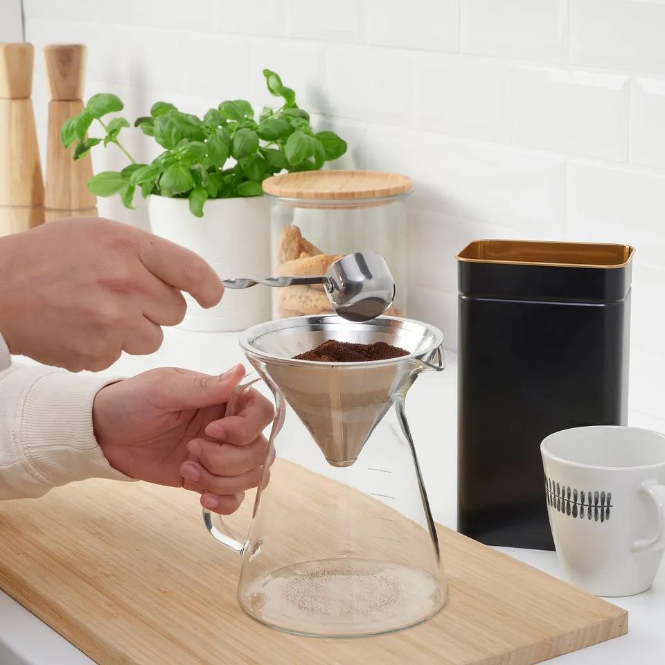 Person pouring coffee grounds into a glass drip philtre on a wooden board. A white mug and a black container are nearby.