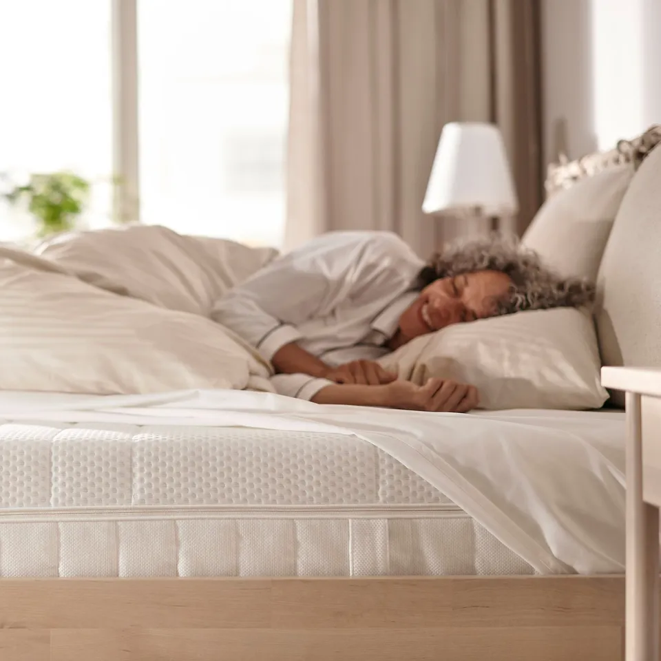 Person sleeping peacefully on white ÅKREHAMN mattress on wooden floor, beige curtains.