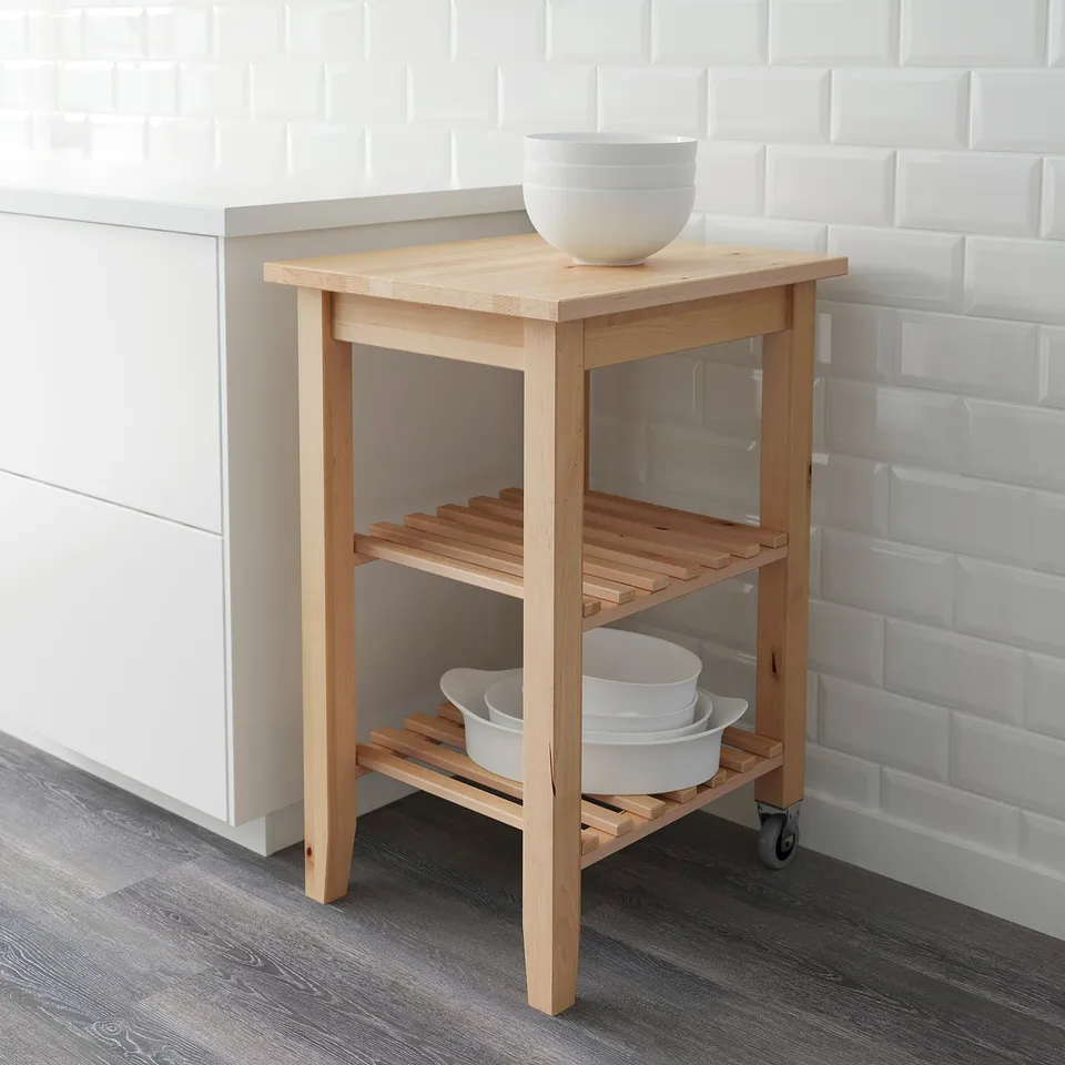 Light wood utility trolley with shelves, holding white dishes against a tile wall.