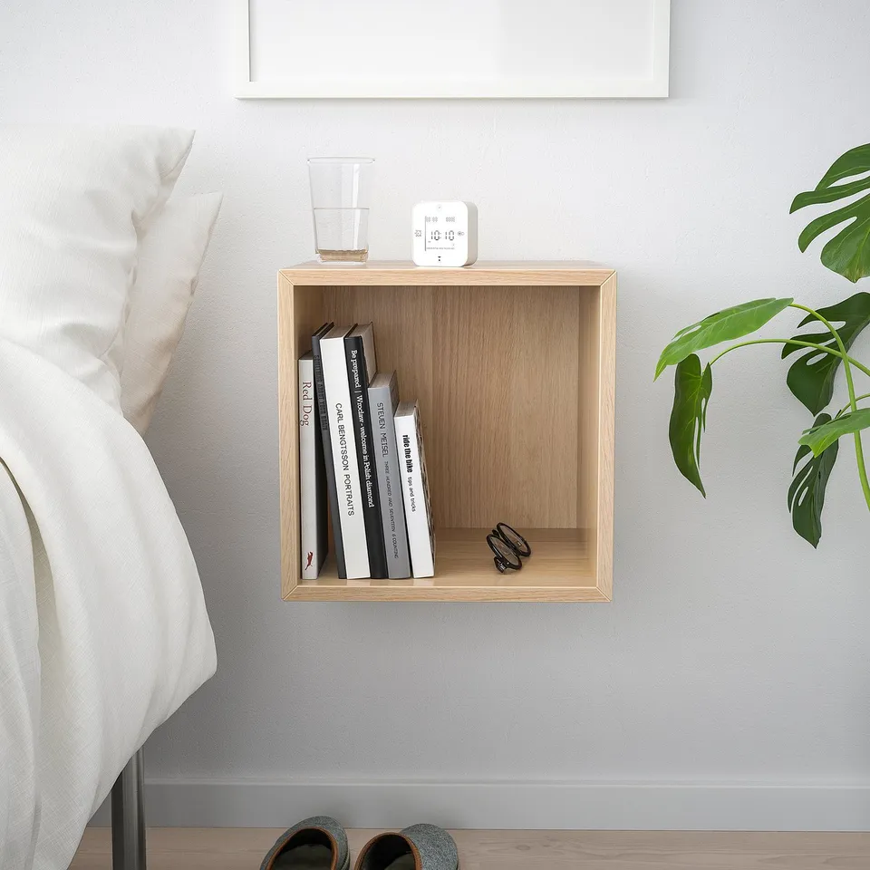 Small wooden wall shelf, white, rectangular shape, holding books, glass, and clock. Next to a bed, with a large leafy plant beside it.