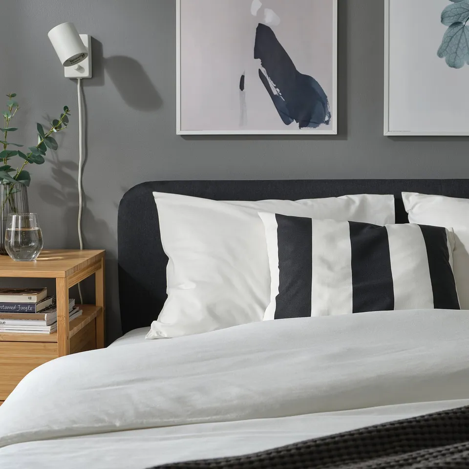 Bedroom with grey wall, black and white striped pillows, art prints, bedside table with books, vase, and lamp.