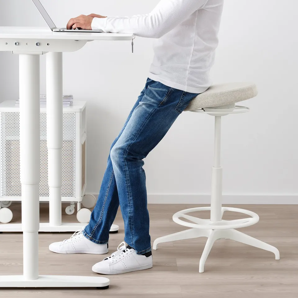 Person sitting on white stool using laptop at tall desk.