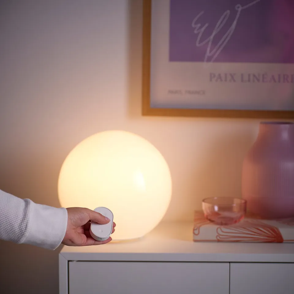 Hand turns off spherical smart lamp on white table. Beside decorative items and framed postcard with paix linéaire.