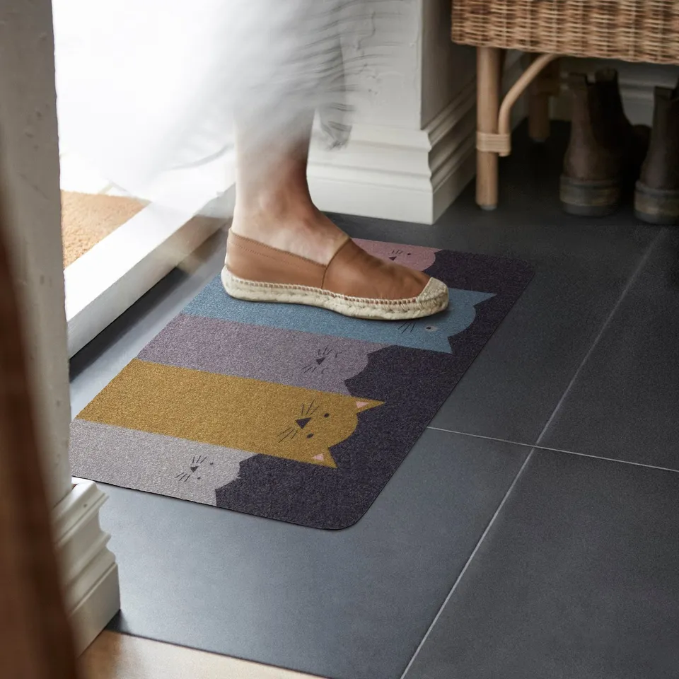 Person stepping on colourful cat TURISTVÄG mat with shoes nearby.