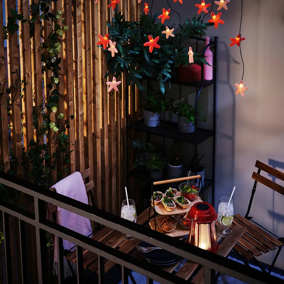 Balcony with TÄRNÖ table and chair, acacia wood, powder-coated steel, lit with star-shaped lights, potted plants, food, drinks.