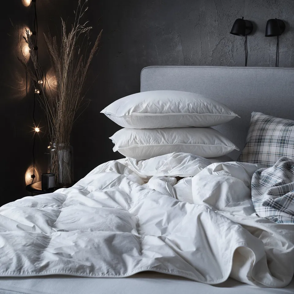 White FJÄLLBRÄCKA duvet on bed with pillows and headboard, grey wall, lights, dried plants.