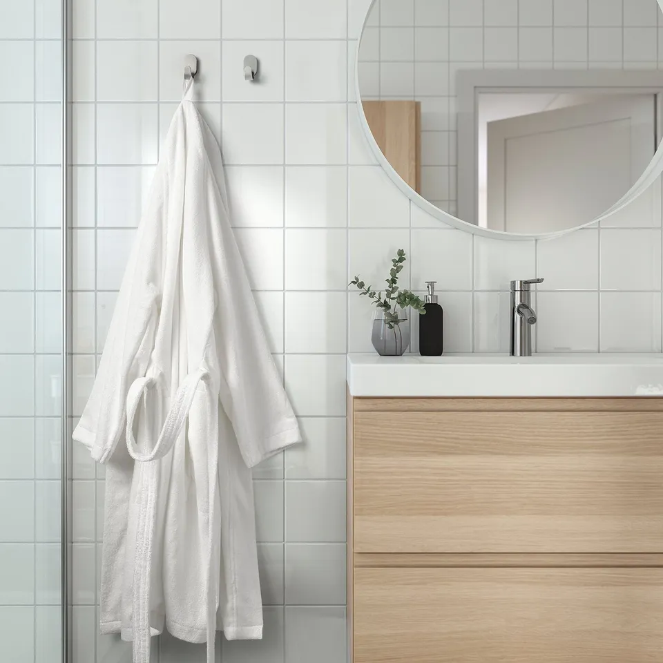 A modern bathroom with white tiled walls. A plush white robe hangs on hooks, beside a wooden vanity with a round mirror and two small vases.