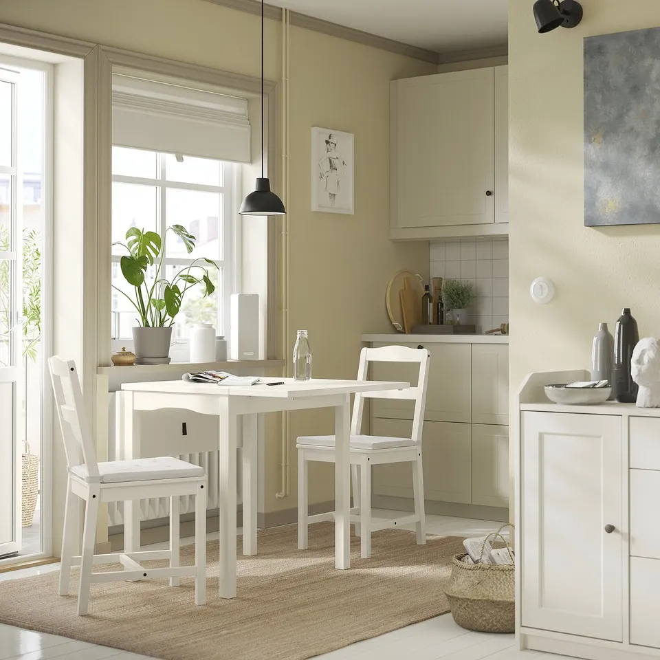 Small cosy kitchen with white JUSTINA chairs, table, and storage cabinet. Green wall, large window, and beige rug. Modern minimalist design.