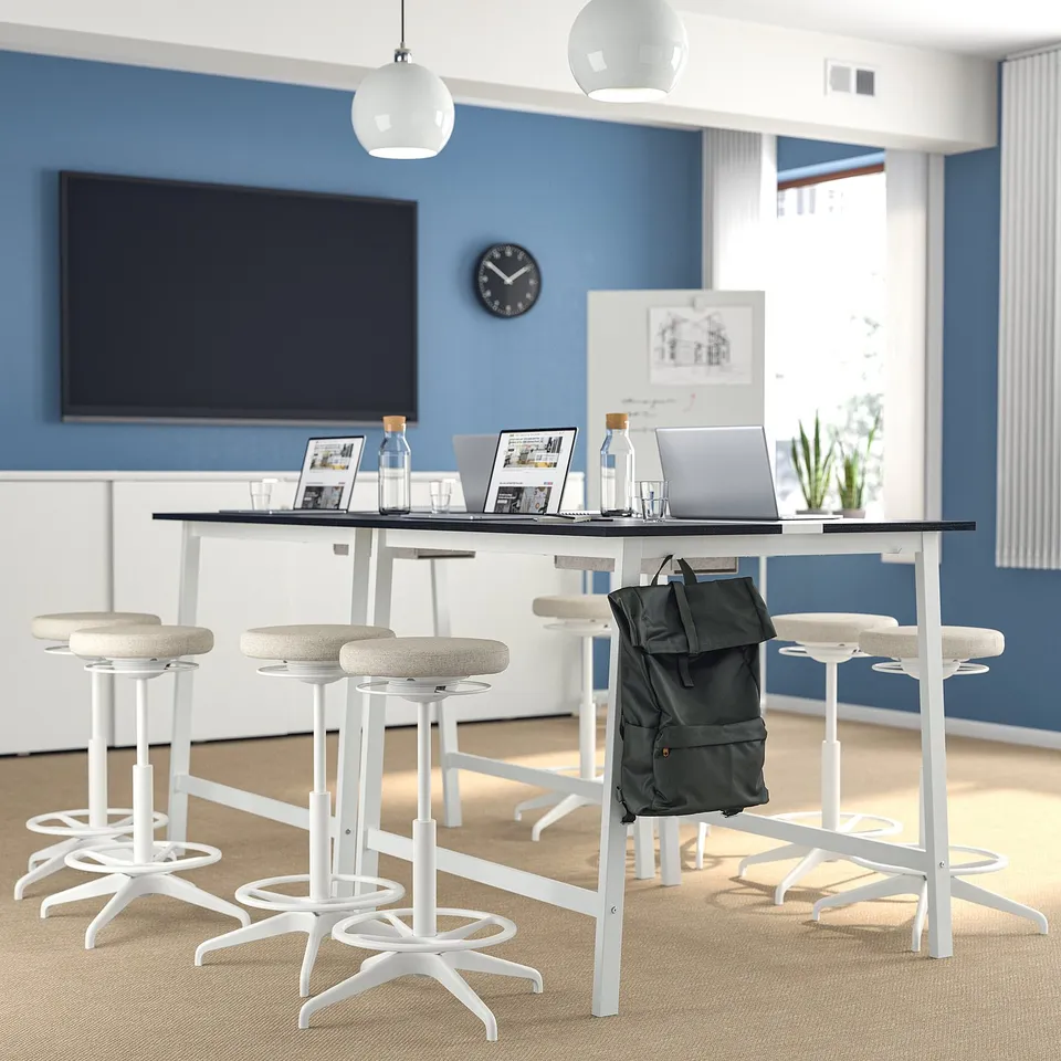 Modern office setup with MITTZON high conference table and white stools. Table has laptops, bottles, and a backpack, with a large tv on the blue wall.