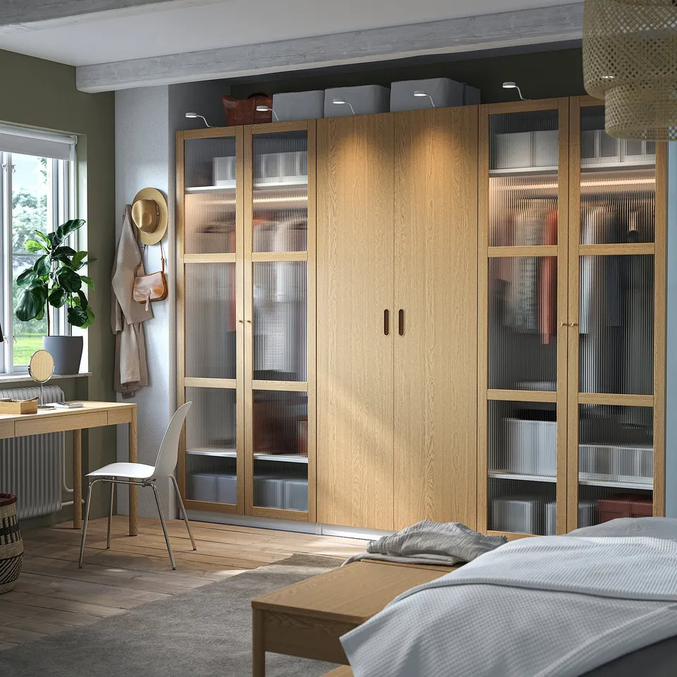Modern room with ikea PAX wardrobe, organized with glass doors. Side desk, plant, decorative items. Bed with rug in foreground, calm lighting.