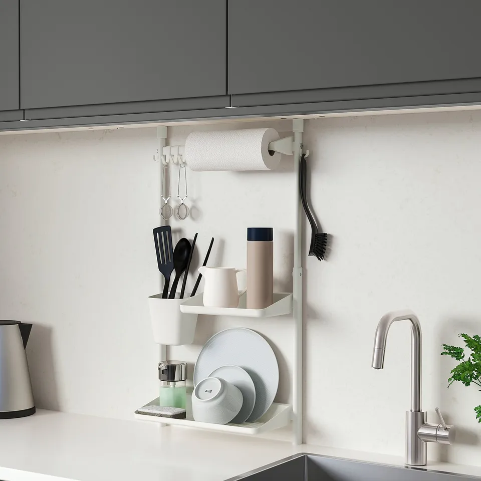 Modern kitchen with SUNNERSTA organiser. White, black utensils, mugs, plates, towel. Silver tap, green plant, grey cabinets.