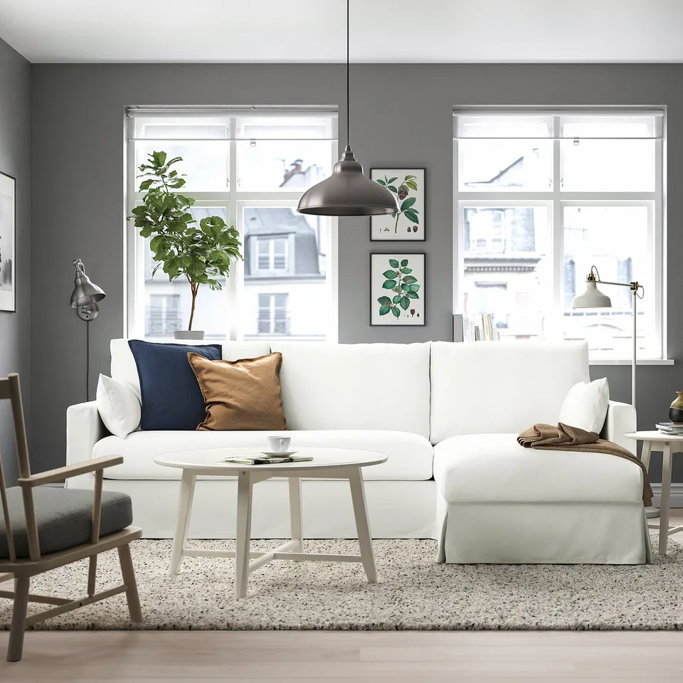 Modern living room with HYLTARP white l-shaped sofa, blue & brown pillows, round white coffee table, grey rug, grey walls, and large windows.