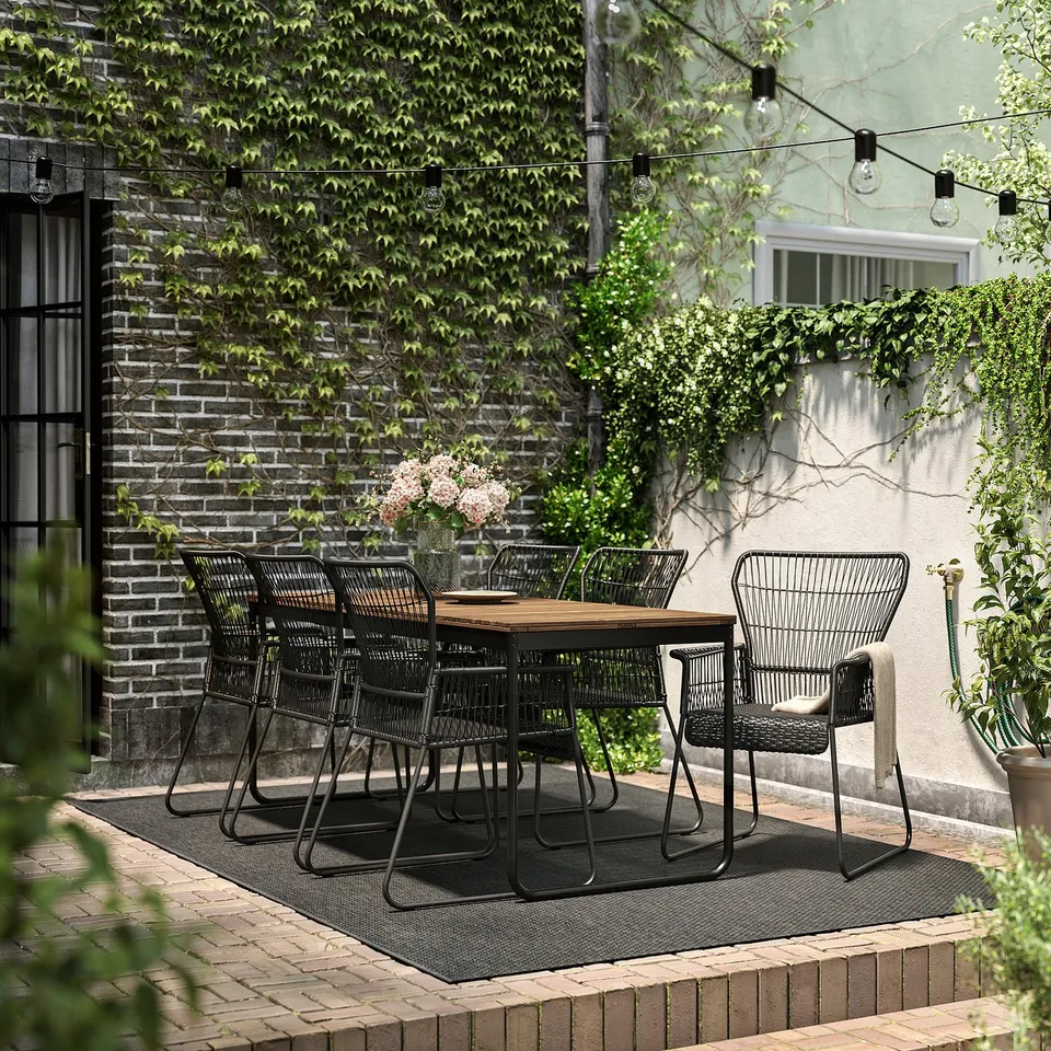 Modern black outdoor dining set with extending table and wicker chairs on a patio, surrounded by plants and string lights.