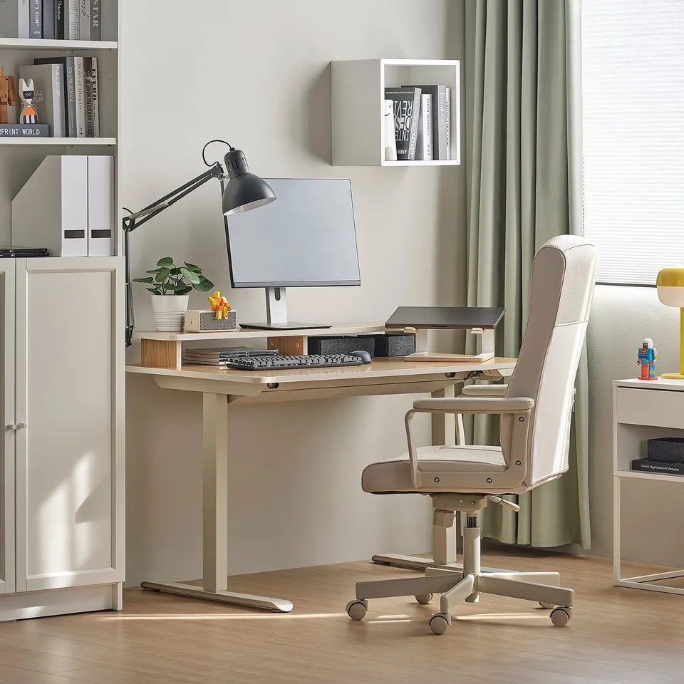 Beige sit/stand desk with black monitor, compact, in corner with office chair, bookshelf, and lamp.
