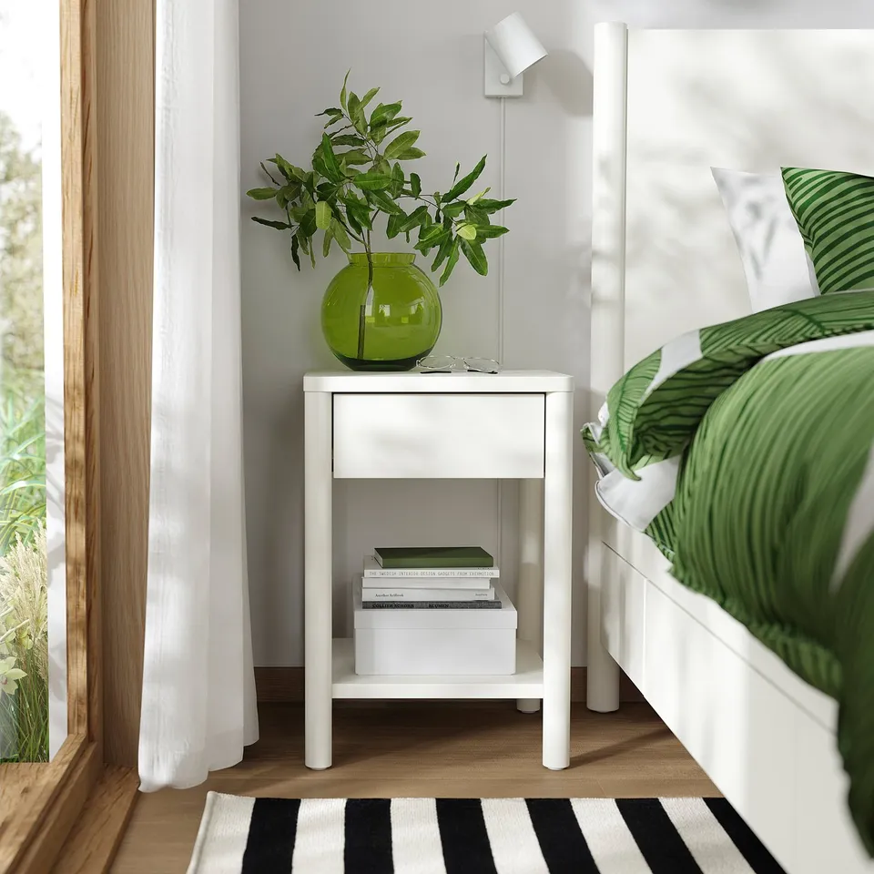 Modern bedroom with white TONSTAD bedside table, plant, books, green-linen bed, striped rug, large window.