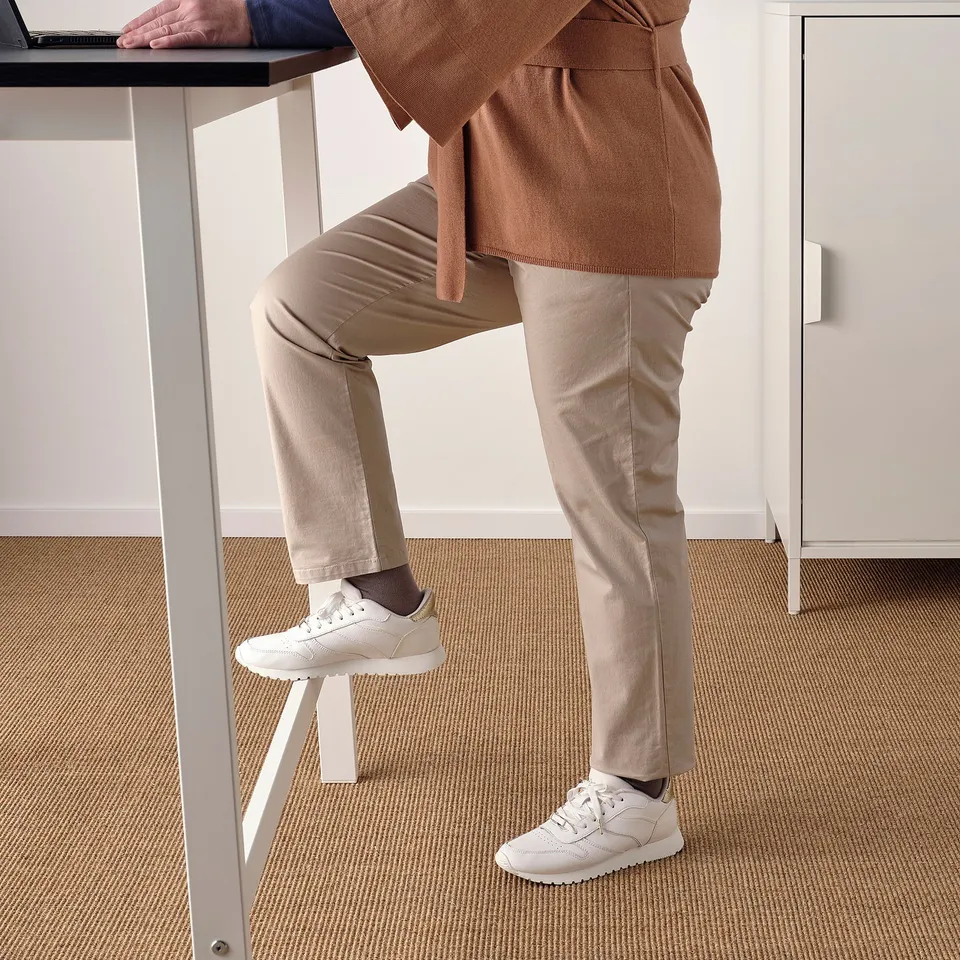 Person leaning on standing desk with feet on stool, wearing white trainers and beige pants.
