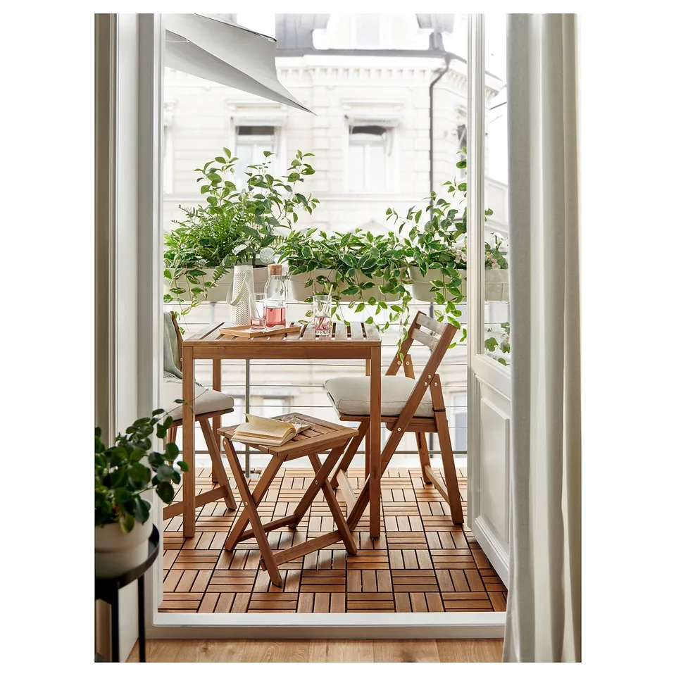 Bright balcony with wooden NÄMMARÖ table and chairs. Lush green plants surround. White building visible through open door.