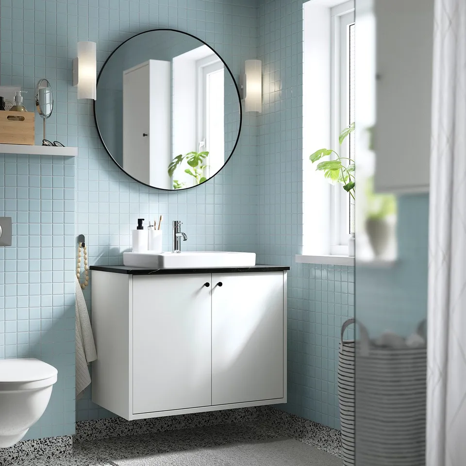 Modern bathroom with HAVBÄCK white cabinet. Round mirror above square sink. Light blue tiles. Plants and towels visible.