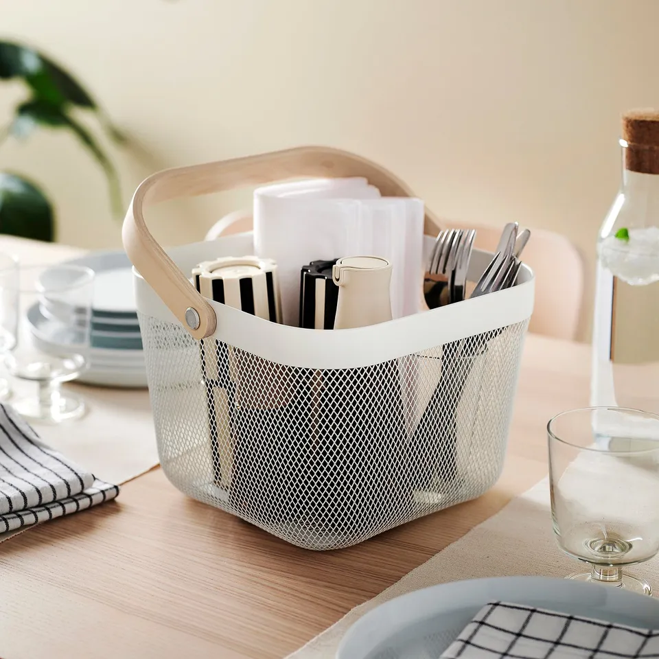 RISATORP storage basket on table, holds cutlery and napkins, made of mesh and birch, small and portable, surrounded by plates and glasses.