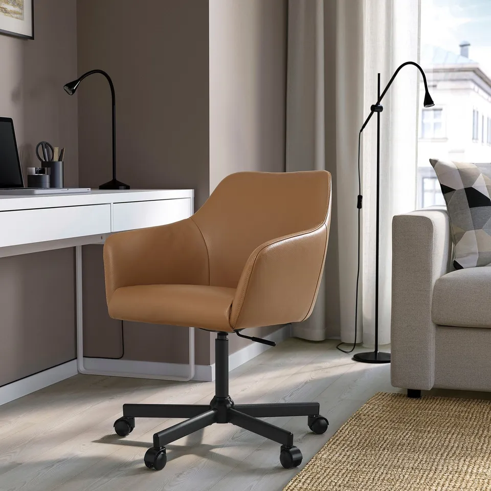 Home office setup: brown TOSSBERG chair, wooden desk, floor lamp, grey sofa, window.