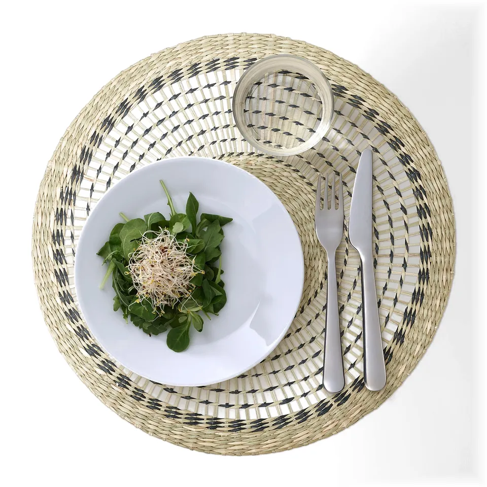 A round woven place mat with a grey and white pattern holds a white plate of salad and cutlery.