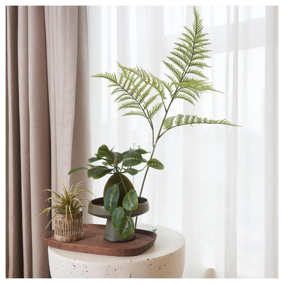 A potted artificial fern with green leaves sits on a white table near a window with sheer curtains.