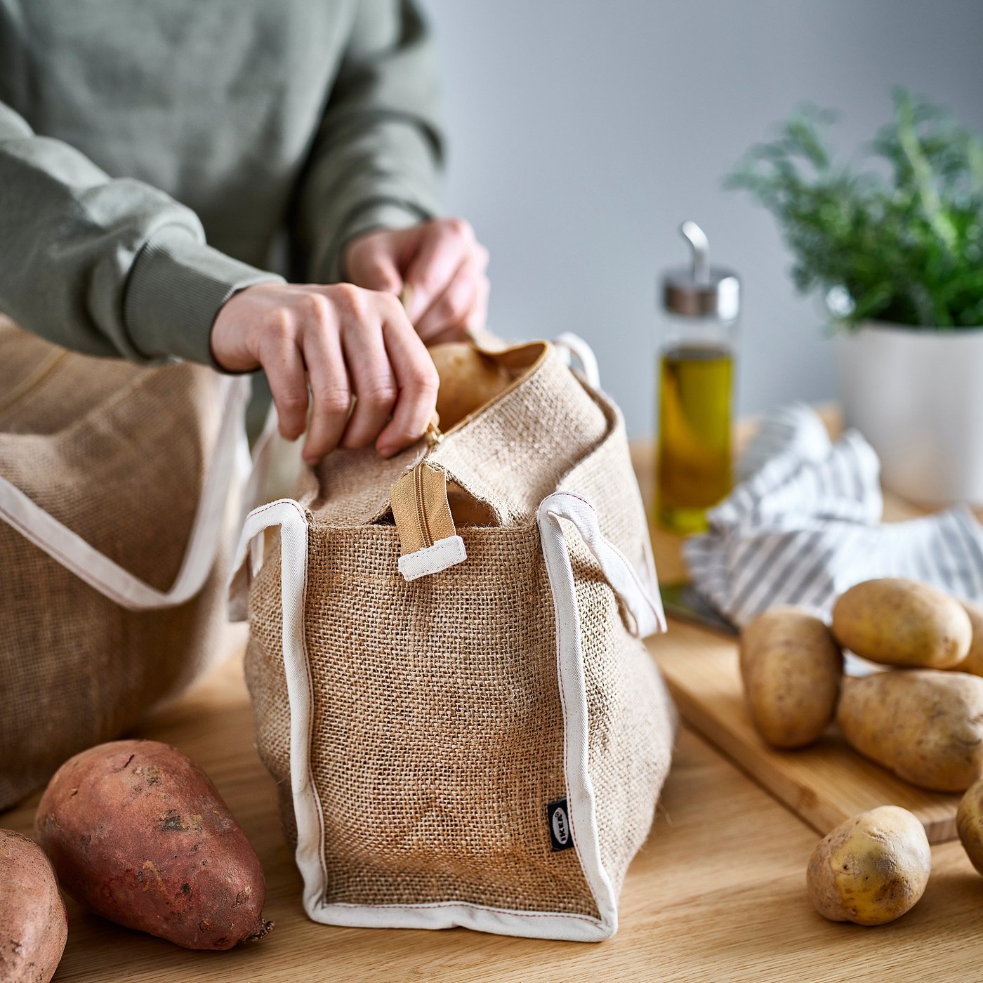 Person opening beige GULLRISMOTT customizable storage bag on wooden table with root vegetables.