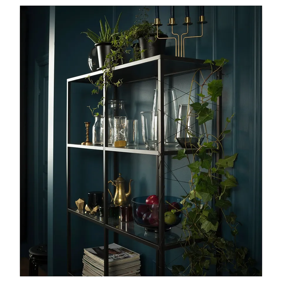 Black-brown VITTSJÖ shelf with glass, holding vases, plants, and fruit bowl against blue wall.