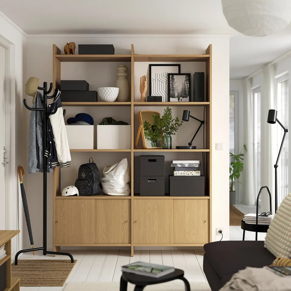 Wooden storage unit, LÅDMAKARE, light beige. Shelves filled with boxes, bags, and decor. Black coat stand at left, black lamp at right.