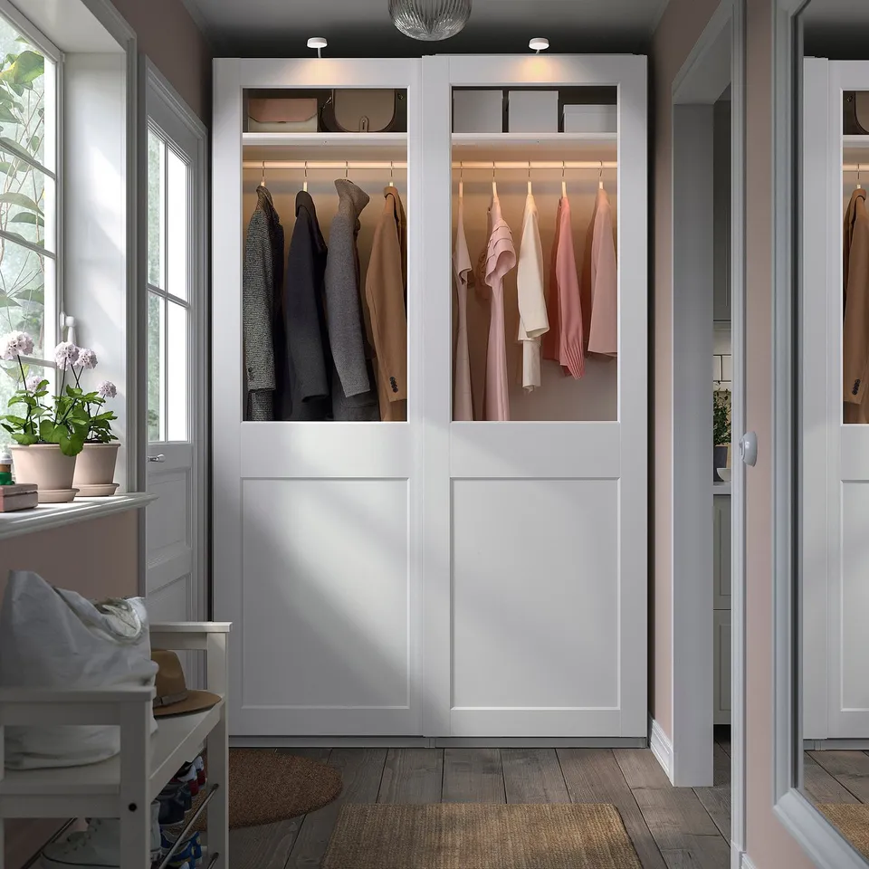 Modern closet with white PAX wardrobes, glass doors, built-in lighting, and wooden stool with towels and a hat.