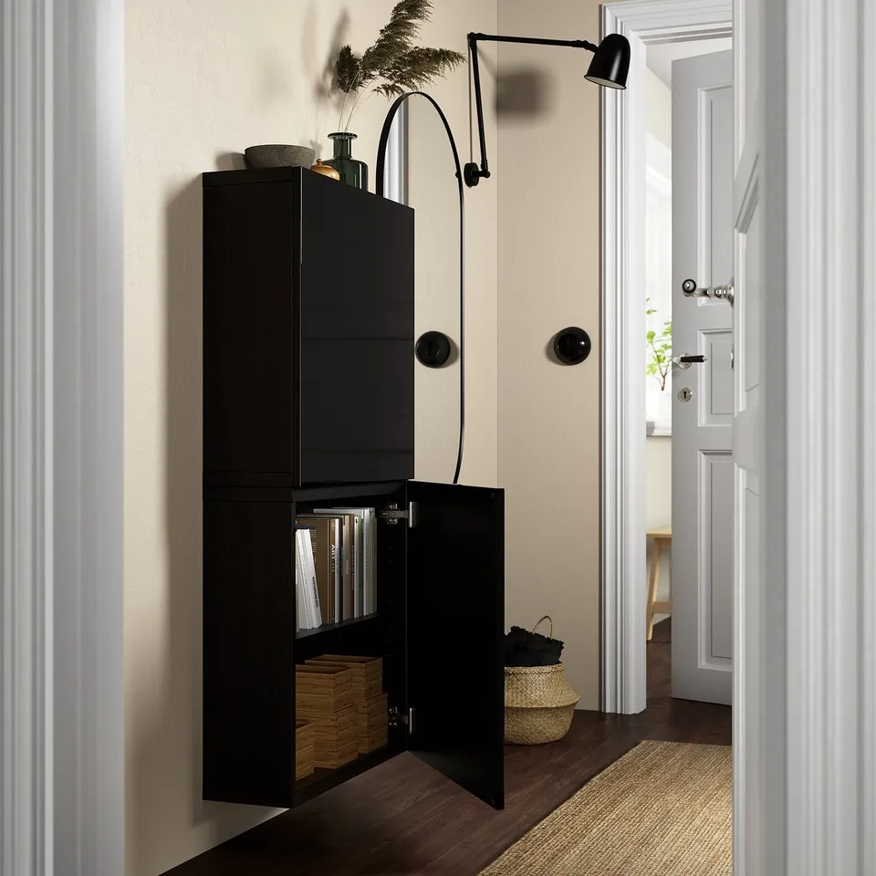 A black wall-mounted storage unit with one door open, revealing shelves with files and woven baskets. A wall lamp and decorative items are on top.