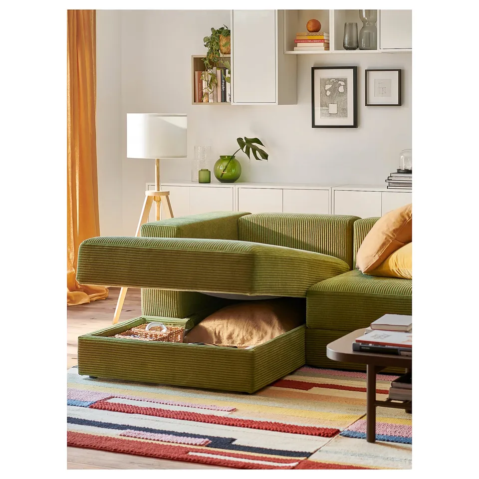 Cosy living room with green JÄTTEBO sofa, cat sleeping under it, shelves with plants, books, and a white lamp.