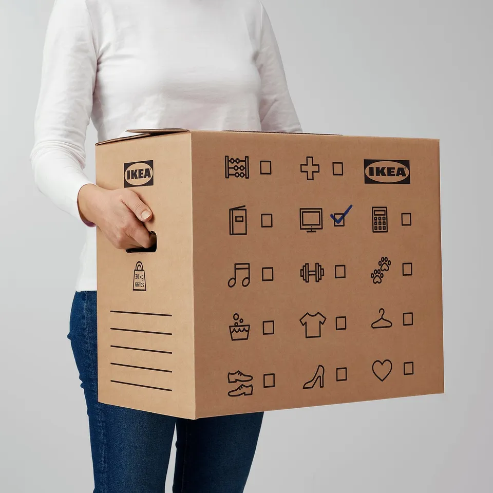 Person holding large cardboard box labelled IKEA with various icons and text indicating contents or categories.