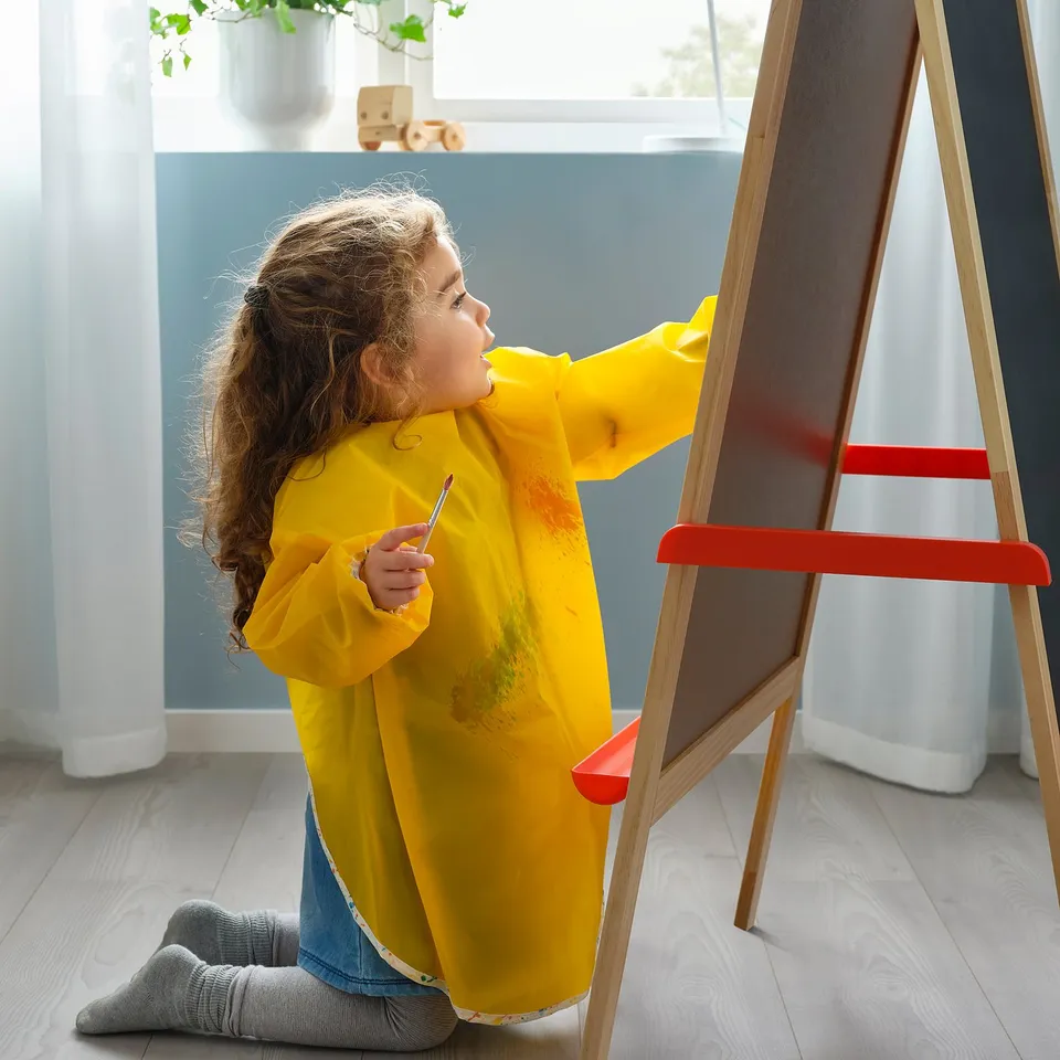 Child wearing yellow MÅLA apron, drawing on easel with red handle, holding paintbrush.