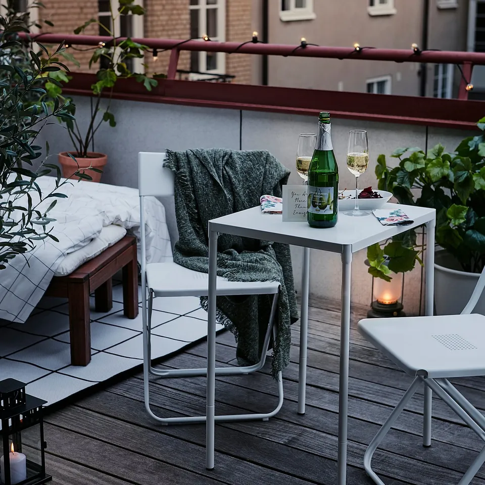 Modern balcony setup with FEJAN white folding chairs, small table, green blanket, and wine glasses. Cosy with plants and lanterns.