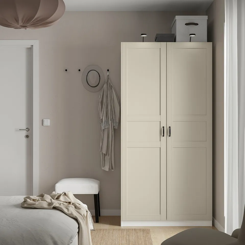 Minimalist bedroom with beige wardrobe, bed, wall mirror, hooks, stool, and YTBERG lamp atop wardrobe.