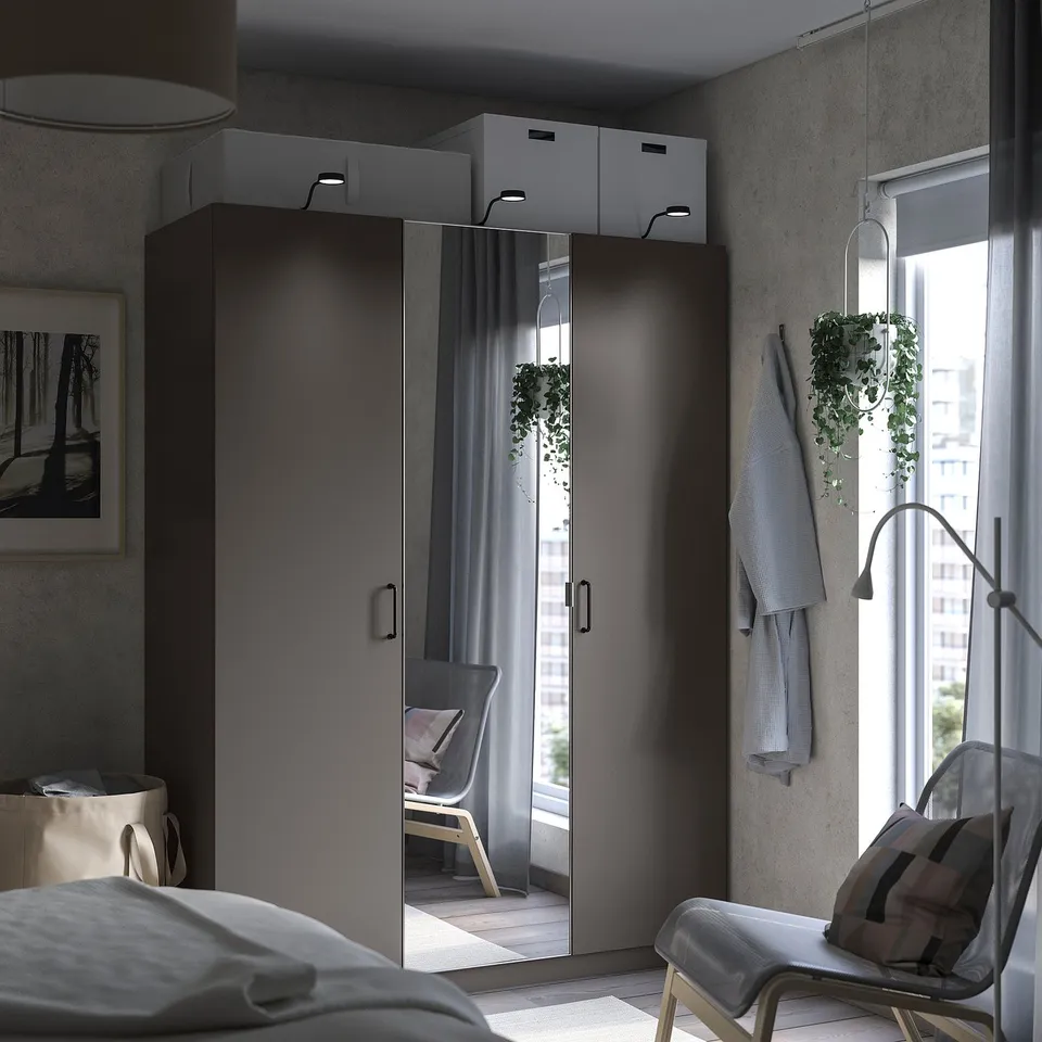 Modern bedroom with grey PAX wardrobe, mirrored doors, white bed, wooden chair, hanging plants, and a window with city view.