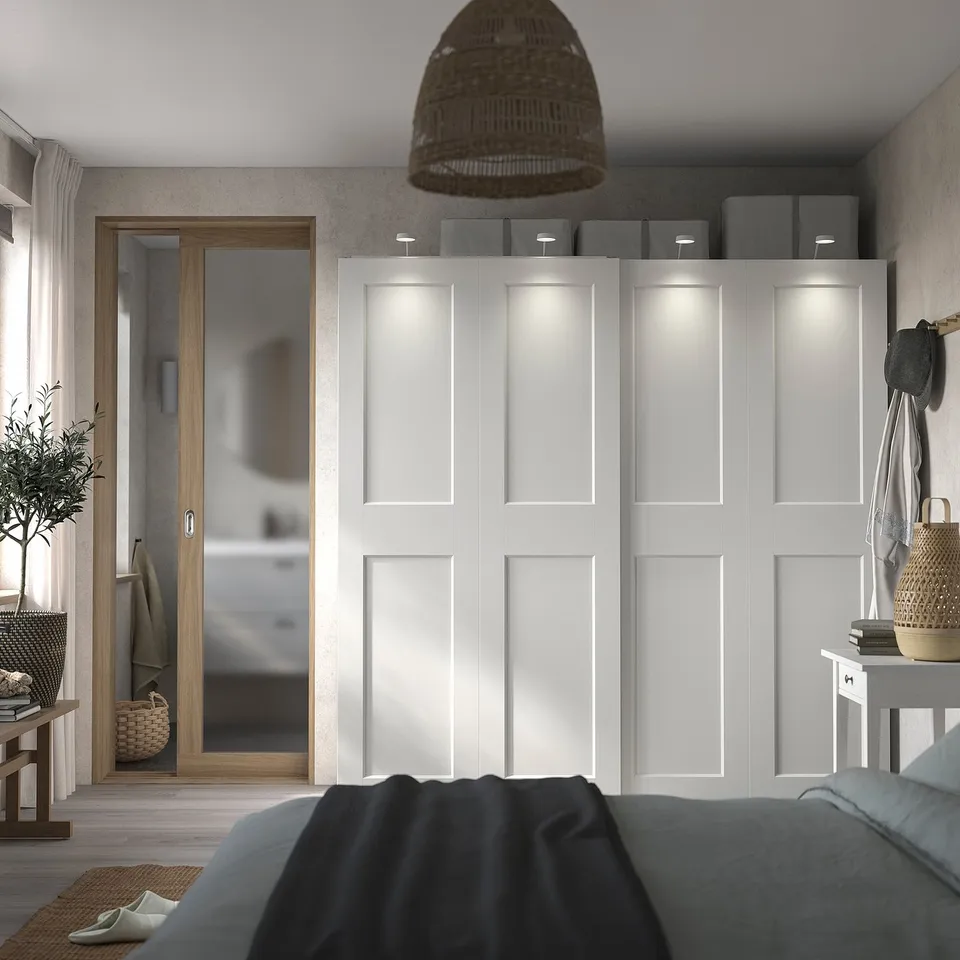 Modern bedroom with white PAX wardrobe, sliding doors, lights, and plants. Grey bed in foreground.