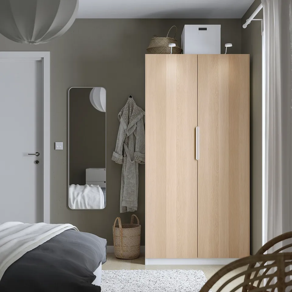 Minimalist bedroom with tall PAX wardrobe, mirror, and wicker baskets. Blue and white bedding, light curtains.