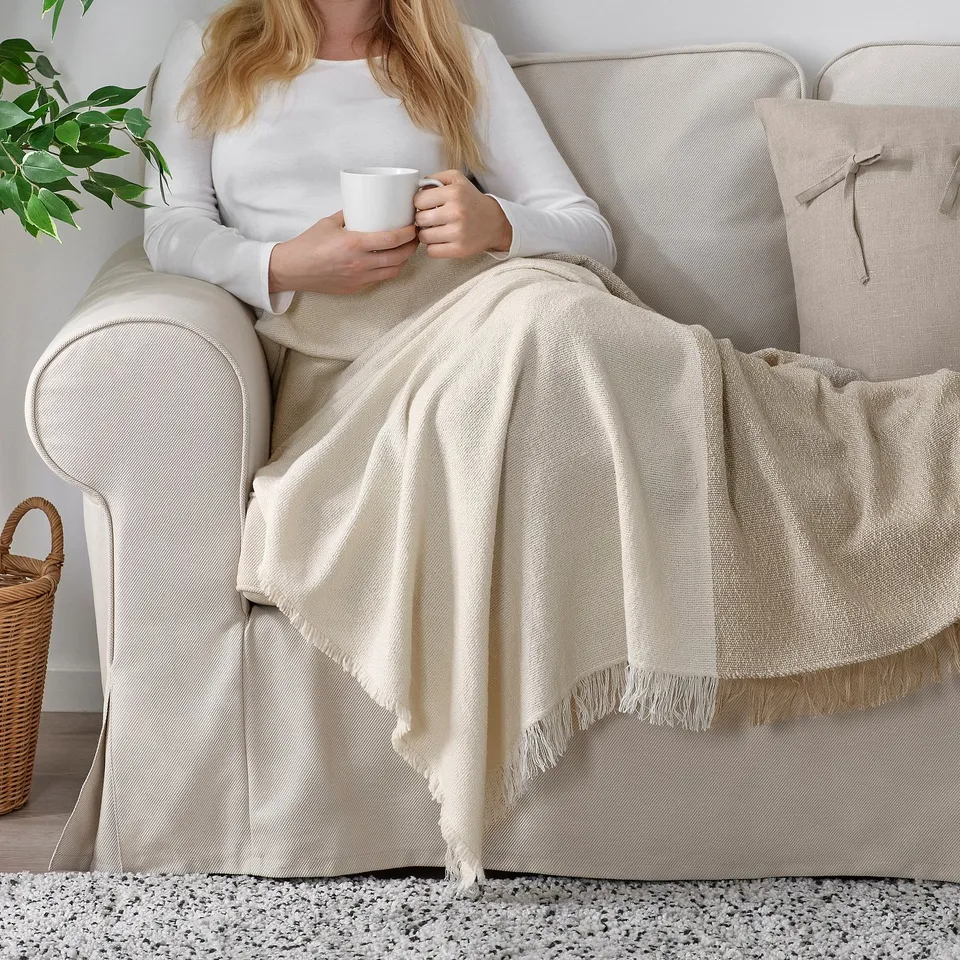 A person sits on a beige couch with a white mug, wrapped in a soft, textured throw blanket with fringes. The blanket is white and grey-beige.