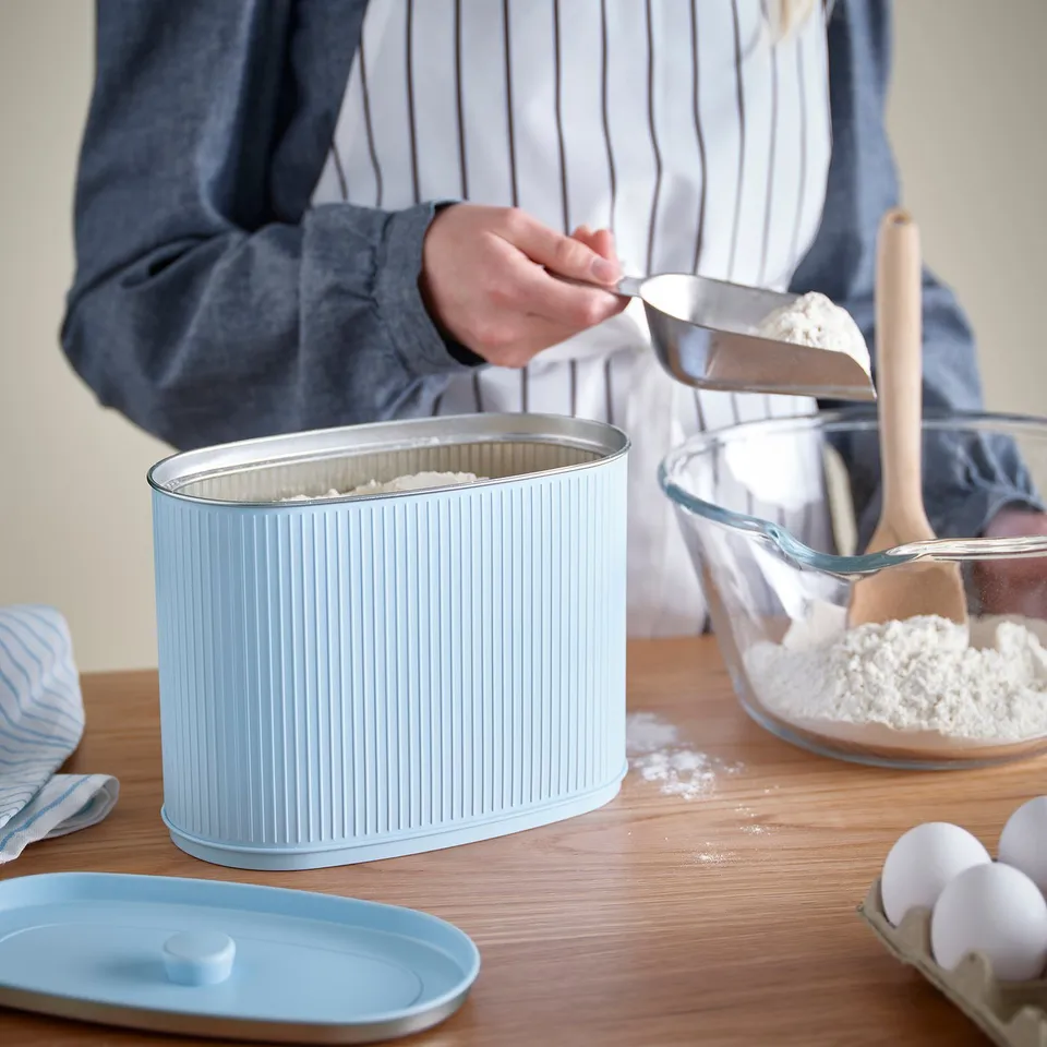 A person scoops flour from a ribbed, light blue storage tin by BETTHAJ onto a wooden table. The tin is surrounded by kitchen tools and ingredients.