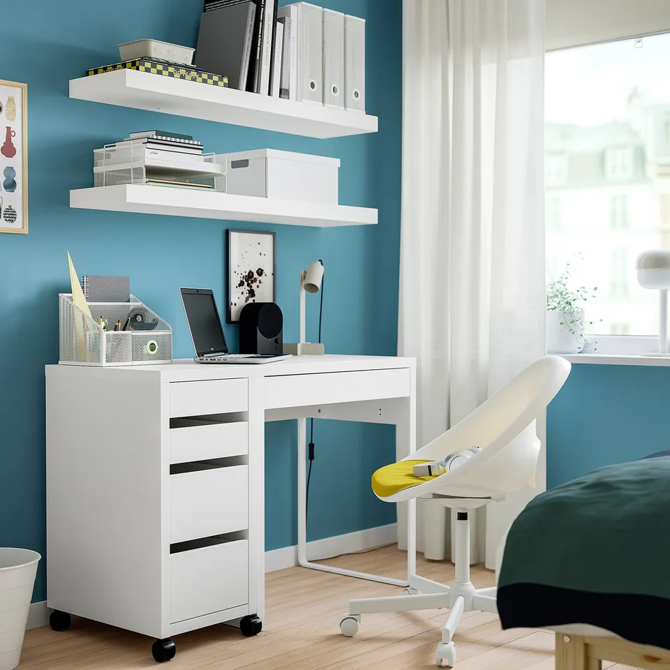 White MICKE desk with drawers and shelves against a blue wall, laptop, and chair.