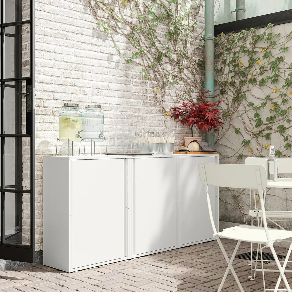 White outdoor cabinet, SUNDSÖ, with adjustable shelves and door, on a brick patio with a brick wall and plants.