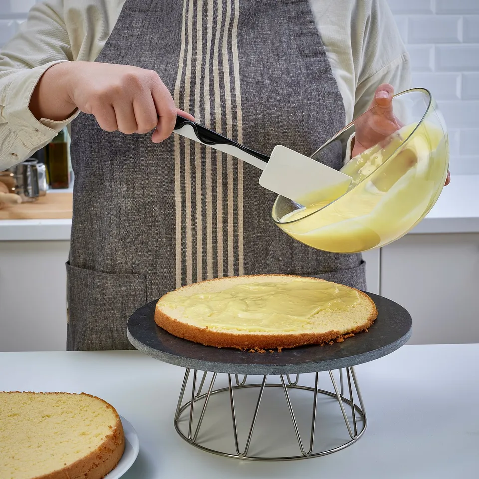 Person spreads yellow icing on cake using a spatula from a glass bowl on a stand.